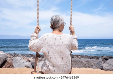 Back view of senior woman sitting on swing on sea beach enjoying relaxed and fun moments, retirement lifestyle for an elderly lady in summer vacation - Powered by Shutterstock