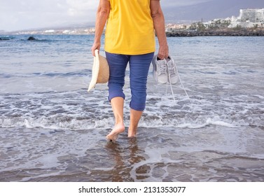 Back View Of Senior Woman Walking Along The Beach With Hat And Shoes In Hands. Mature Lady Bare Feet In The Water All Dressed Up. Scenic Nature And Horizon In The Background