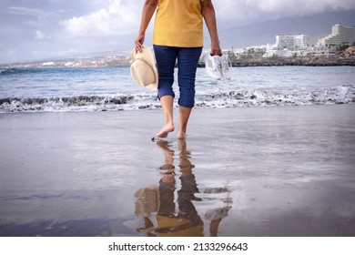 Back View Of Senior Woman Walking Along The Beach With Hat And Shoes In Hands. Mature Lady Bare Feet In The Water All Dressed Up. Scenic Nature And Horizon In The Background