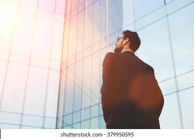 Back view of serious businessman in eye glasses looking on copy space while standing against glass skyscraper, young professional employee waiting for international partners outdoors near big company