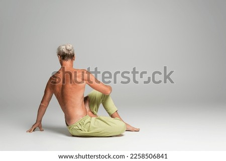 back view of shirtless man doing supine spinal twist yoga pose on grey background