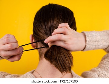 Back View Shot Of A Fair-haired Lady. There's A Black Rubber Band On Her Hair With A Black Fake Hair Braid. The Photo Was Taken Against A Yellow Background.