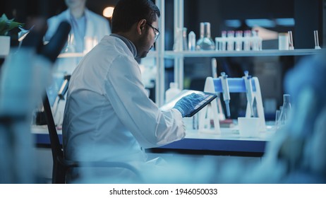 Back View Shot An Indian Male Working In A Modern Applied Science Laboratory. Using Digital Tablet Computer To Enter Analysis Daa After Successful Biological Experiment. Dark Lab With Cold Color.