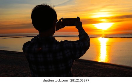 Back view with Silhouette of a man capturing a golden sunset reflection on the beach using a mobile phone a stunning ocean sunset during vacation