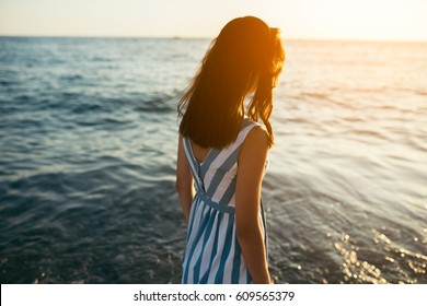 Back view silhouette of portrait brunette beautiful woman walking along beach and sea sunset background. Cover idea. Female in dress walking on ocean look to the sun. Loneliness and sadness concept.