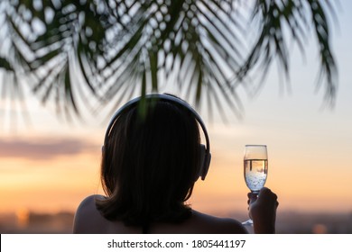 Back view silhouette of relaxed woman with glass of champagn and headphones listening to music on the beach at sunset in the branches of palm trees.  - Powered by Shutterstock