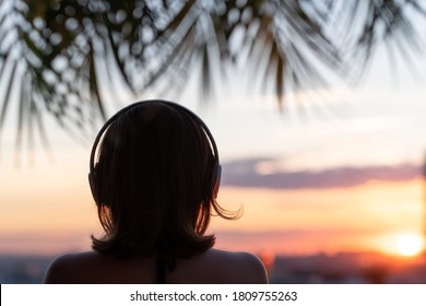 Back view silhouette of relaxed woman wearing headphones meditating listening to music on the beach at sunset in the branches of palm trees.   - Powered by Shutterstock