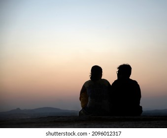 Back view of silhouette traveler couple sitting together and looking at beautiful sunrise or sunset on the top of mountain with clear sky in background. Man and woman watch the sunrise morning