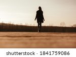 Back view silhouette of unrecognizable female in warm clothes walking alone on dry grassy meadow in sunny autumn evening in countryside