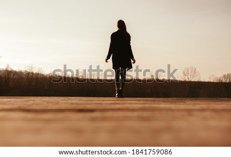 Back view silhouette of unrecognizable female in warm clothes walking alone on dry grassy meadow in sunny autumn evening in countryside