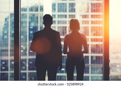 Back View Silhouettes Of Two Business Partners Looking Thoughtfully Out Of A Office Window In Situation Of Bankruptcy,team Of Businesspeople In Fear Or Risk Watching Cityscape From Skyscraper Interior