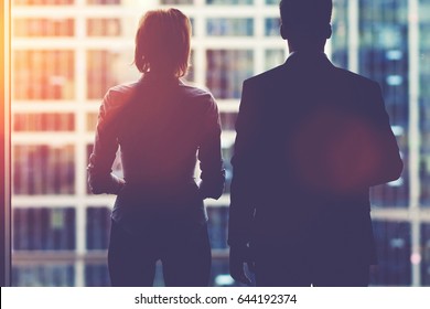 Back View Silhouettes Of Two Business Partners Looking Thoughtfully Out Of A Office Window In Situation Of Bankruptcy,team Of Businesspeople In Fear Or Risk Watching Cityscape From Skyscraper Interior