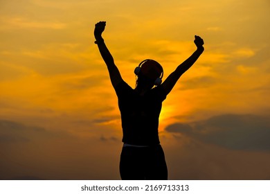Back view silhoutte sport woman wearing headphone and hands up watching sunset, silhouette slender sporty female working out exercising in the evening, fitness woman warm up after doing exercise