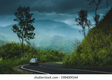 Back View Of Silver SUV Car Driving On The Curve Mountain Road. Dramatic Rain Clouds Covers Mountains And Forest In The Backgrounds. Exploration, Travel, Vacations Concepts. Focus On The Car.