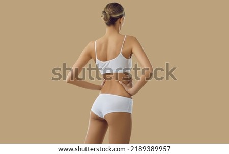 Back view of slender female model posing in white underwear. Backview of young European woman with slim skinny body wearing basic soft natural cotton lingerie set standing on beige color background