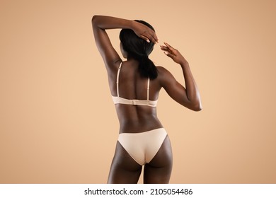 Back view of slim dark skinned african american woman in beige underwear demonstrating her perfect body, posing against beige studio background with hands up, copy space, slimming concept