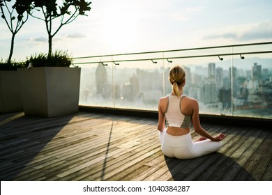 Back view of slim female in active wear sitting in lotus pose recreating on spiritual practice and keeping healthy lifestyle, caucasian female relaxing on yoga practice feeling calm on rooftop terrace - Powered by Shutterstock