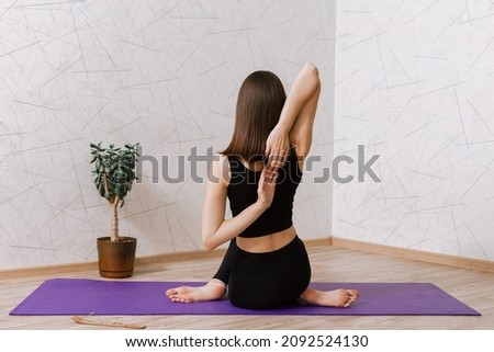 Back view of slim flexible woman practicing yoga in Cow Face Gomukhasana pose while sitting on mat in room at home 