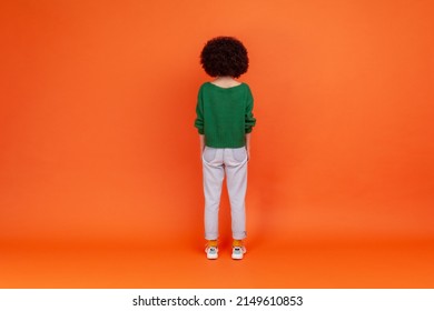 Back view of slim woman with Afro hairstyle wearing green casual style sweater and jeans standing and looking at something, waiting. Indoor studio shot isolated on orange background.
