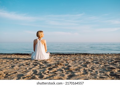 Back view of slim woman with short fair hair wearing white light summer sundress sitting on sandy beach near water sea ocean on sunny day, enjoying sunshine. Summer, holiday, travelling, vacation.