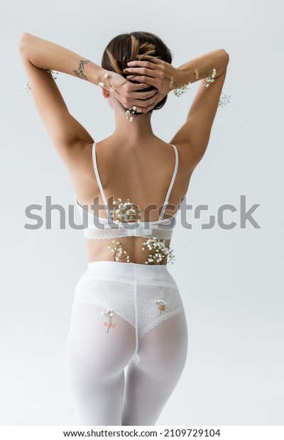 back\
view of slim woman in white bra and tights, with tiny flowers on\
body, posing with hands behind head isolated on\
grey