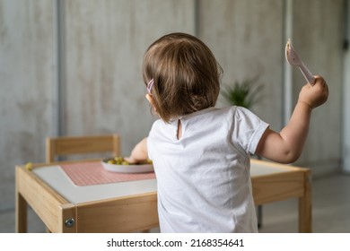 Back View Of Small Caucasian Girl Little Child Toddler Holding Plastic Fork While Reaching For The Food On The Table At Home In Room Alone Real People Copy Space