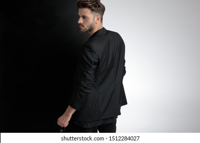 Back View Of Smart Casual Man Holding A Pair Of Sunglasses, Looking To Side And Standing On Black And White Background In Studio