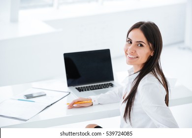 Back view of smiling business woman sitting by the table with laptop and looking at camera in office