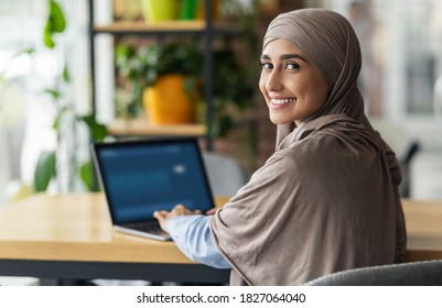 Back view of smiling muslim lady in hijab working or studying online at cafe, using laptop, copy space. Happy arab woman in headscarf freelancer typing on laptop, enjoying her distant job