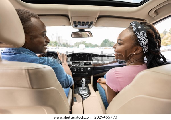Back view of smiling young black lovers man and\
woman in casual comfortable outfit looking at each other, excited\
afro american couple having car trip by comfy luxury auto, shot\
from backseat
