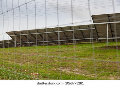 Back view of solar panels set at angle in green and brown autumn field behind wire fence under grey sky in rainy day in Portugal (FOCUS ON RIGHT PANEL)