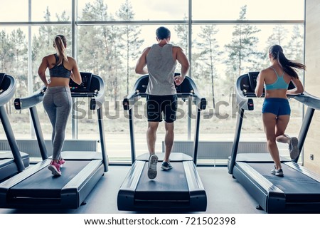Back view of sports people on running track. Two attractive young women and handsome muscular man are running in gym. Treadmills.