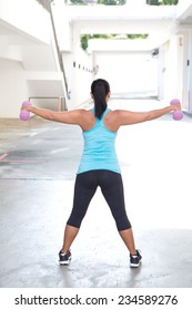 Back View Of Sporty Woman Holding Pink Barbell With Both Arms Stretched Out For Shoulder Strengthening, Outdoors.