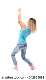 Back View Of Standing Girl Pulling A Rope From The Top Or Cling To Something. Girl  Watching.   Isolated Over White Background. Blonde In A Blue Shirt And Jeans, Pulls The Top Of The Rope.