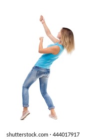 Back View Of Standing Girl Pulling A Rope From The Top Or Cling To Something. Backside View Of Person. Blonde In A Blue Shirt And Jeans, He Pulls The Top Rope For Turning Right Side.