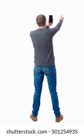 Back View Of Standing Young Man  And Using A Mobile Phone. Girl Watching. Rear View People Collection. Isolated Over White Background. A Guy In A Gray Sweater Photographs Smartphone And Shows Thumb Up