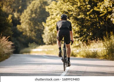 Back View Of Strong Man In Sport Clothing And Black Helmet Riding Professional Bike On Nature. Mature Sportsman Enjoying Favorite Hobby On Paved Road.