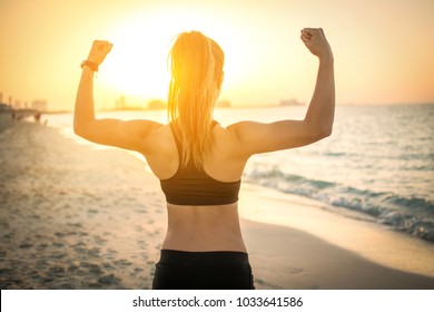 Back view of strong sporty girl showing muscles at the beach during sunset.