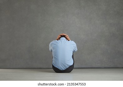 Back view studio portrait of man sitting on floor facing grey stone wall, holding head, trying to hide true emotions, feeling desperate, distressed, frustrated, hopeless, unable to cope with problem