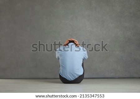 Back view studio portrait of man sitting on floor facing grey stone wall, holding head, trying to hide true emotions, feeling desperate, distressed, frustrated, hopeless, unable to cope with problem