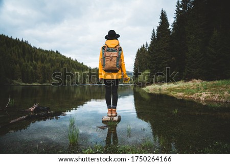 Back view of stylish hipster woman holding flowers,wearing vintage backpack, hat and yellow jacket looking at mountain view while relaxing in nature. Travel and wanderlust concept.Amazing chill moment