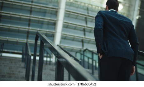 Back view successful man walking alone at city center. Confident businessman going upstairs in luxury suit near stadium. Male entrepreneur putting hand in pocket at stairs outdoors.