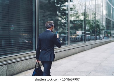 Back View Of Successful Middle Aged Man In Stylish Dark Business Suit Walking Down City Street With Smartphone In Hand Near Business Center In New York