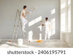 Back view of a team of professional painters in white clothes painting a grey wall in empty room with paint roller. Building contractors doing repair renovation in client house. Repair concept.