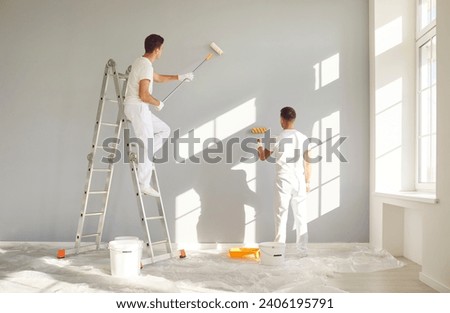 Back view of a team of professional painters in white clothes painting a grey wall in empty room with paint roller. Building contractors doing repair renovation in client house. Repair concept.