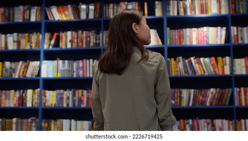 Back View Of Teen Girl Looking At Bookshelves In Library. Schoolgirl In Modern Bookshop Selecting Book To Buy Standing In Front Of Bookcase. Education And Hobby Concept