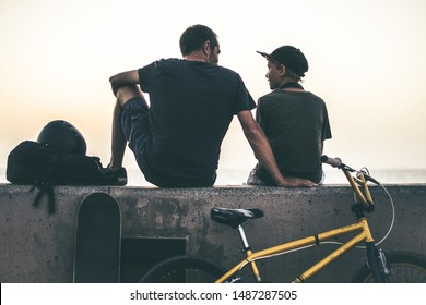 Back view of a teen and his father sitting on a wall with bicycle and skateboard. Young rider and dad relaxing after a fun day of jump and tricks with bmx People waiting friends to go to the skatepark - Powered by Shutterstock