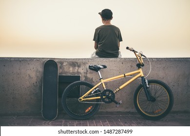 Back View Of A Teen Sitting On A Wall With Bicycle And Skateboard. Young Rider Relaxing After A Fun Day Of Jump And Tricks With His Bmx. Teenager Waiting Friends For Going To The Skatepark Together.