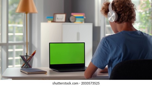 Back View Of Teenage Student In Headphones Writing Notes During Web Lesson On Laptop At Home. Green Screen. Rear View Of Teen Boy Do Homework Or Study Remotely