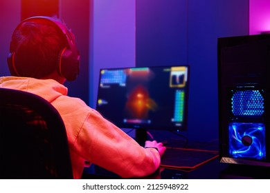 Back view of teenager boy play computer video game in dark room, use neon colored rgb mechanical keyboard, place for cybersport gaming, soft focus on boy